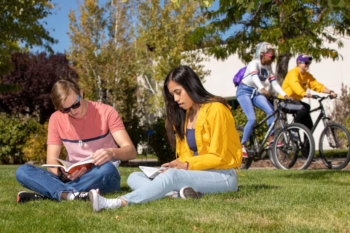 Two students riding bikes and two students studying while sitting on lawn.