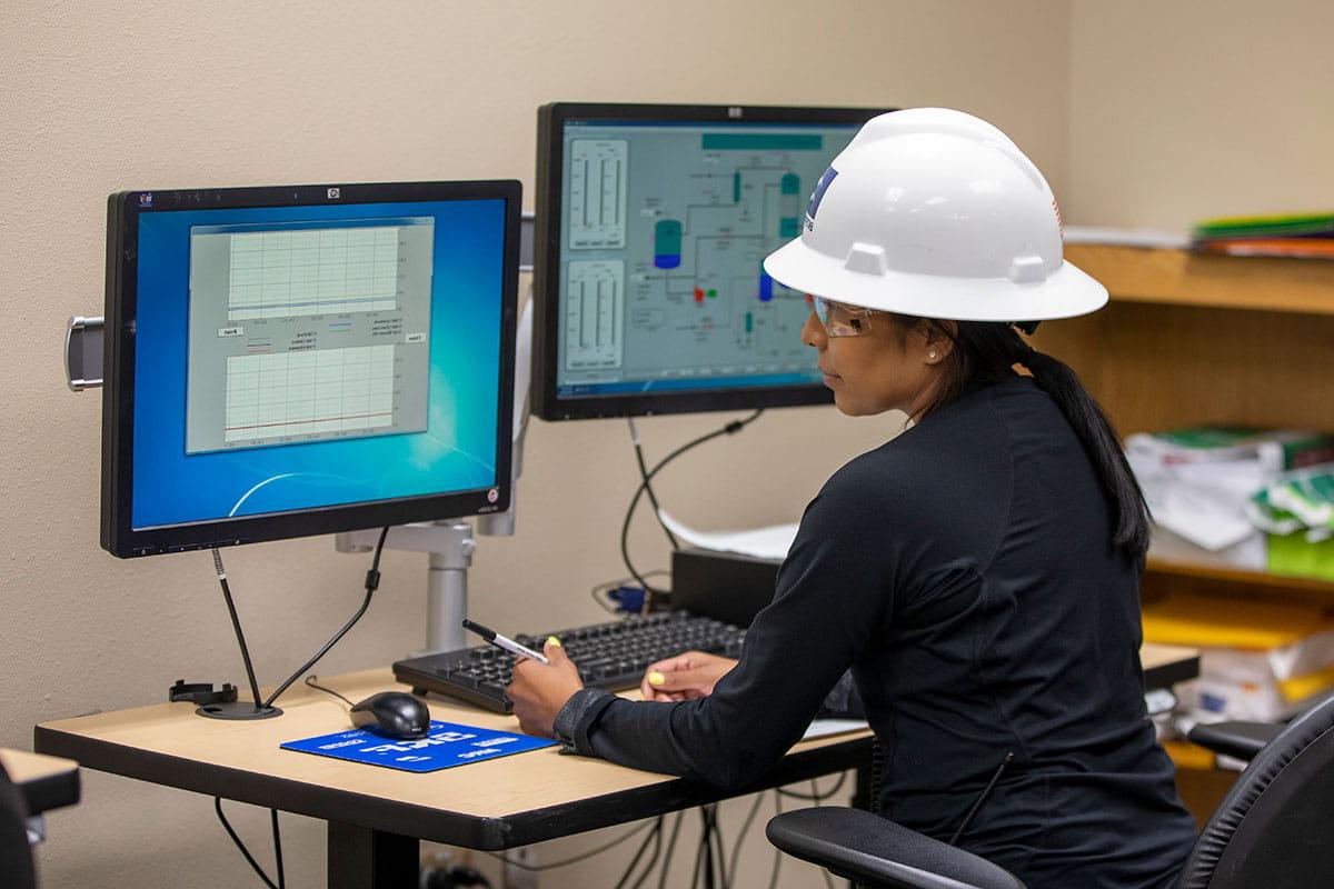 An individual in a white hard hat and safety goggles sits at a desk looking at a computer screen