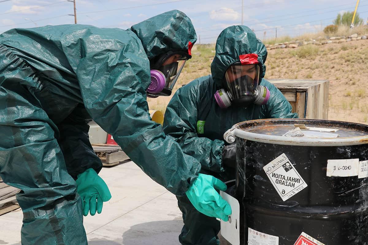 Two individuals in hazmat suits inspect a container of hazardous materials