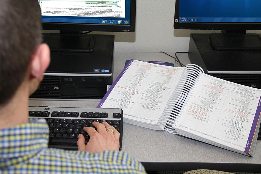 一个人坐在电脑前, 在键盘上打字, 他们旁边的桌子上放着一本医学编码信息的书