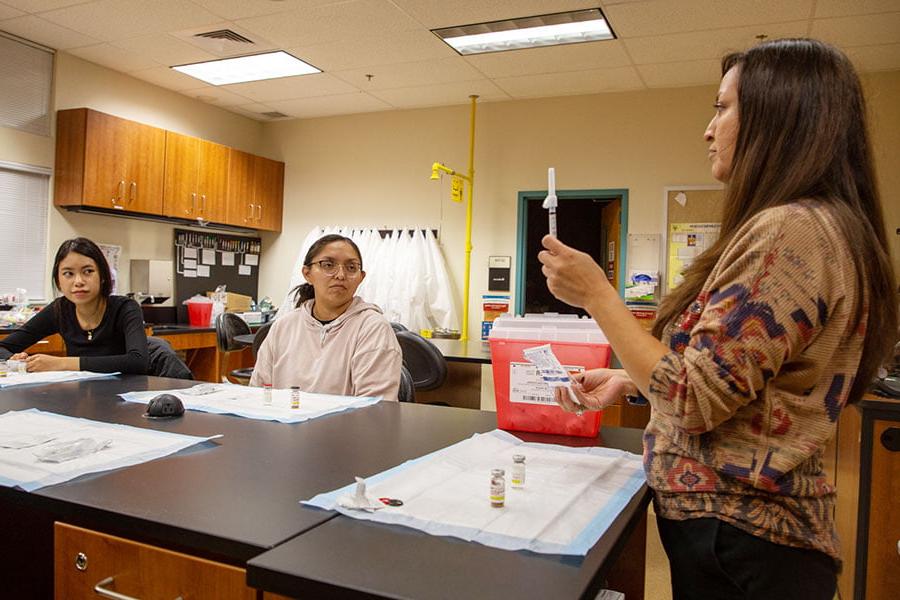 一名教师站在桌子前拿着医用注射器，两名学生在旁边看着